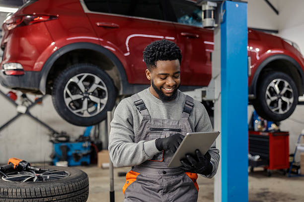 Smiling multicultural repairman standing at car service workshop with tablet in hands and scrolling on it.