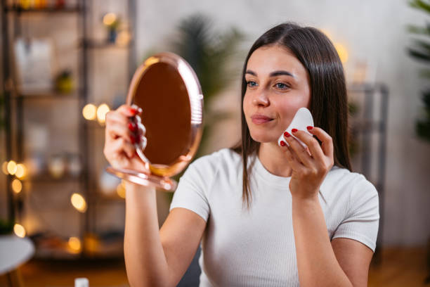 istock Young Woman Using A Gua Sha Stone While Looking In A Hand Mirror In The Living Room 2201019182