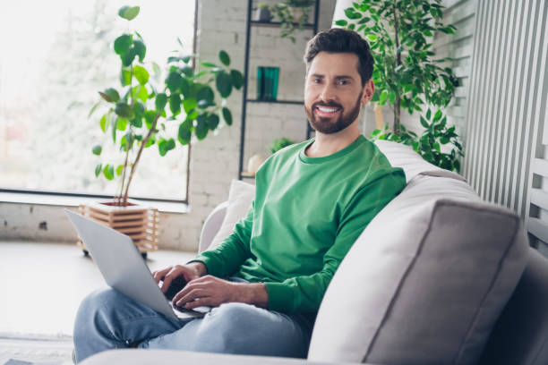 istock Smiling man sitting on a sofa at home working on a laptop, enjoying a relaxed indoor setting with natural light 2201021689