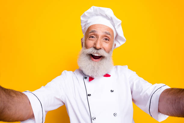 istock Happy senior chef with cheerful expression posing in kitchen uniform against bright yellow background 2201023025