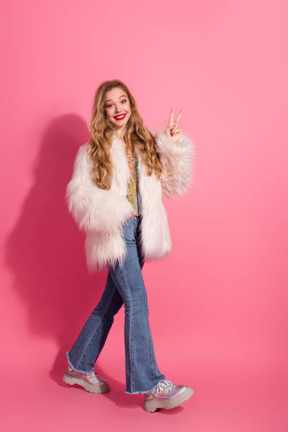 istock Stylish young woman in a fur coat posing with a cheerful expression and making a peace gesture against a vibrant pink vertical background 2201026101