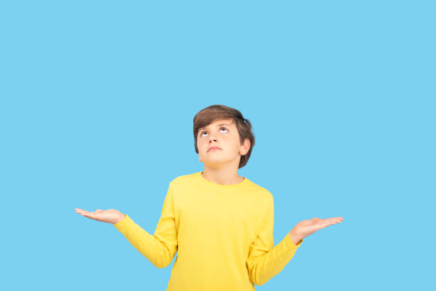 istock Confused young boy shrugging with both hands, looking up with uncertain expression, wearing yellow sweater, isolated on blue background, thinking, doubt, decision 2201033311