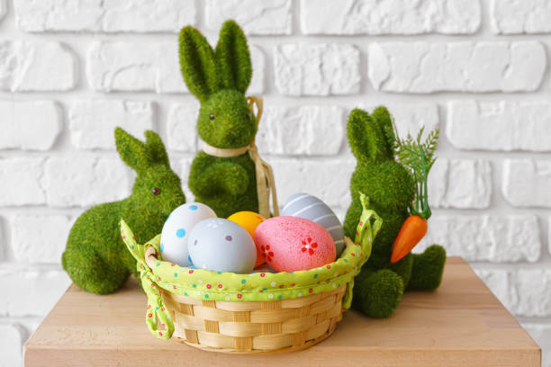 Decorative green moss bunny figures with Easter eggs in wicker basket on white brick wall background