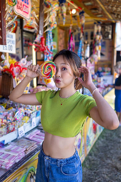 Three-quarter-length portrait of a young adult female having fun at a fairground in Newcastle-upon-Tyne, North East England. She is holding a multicoloured giant lollipop in front of one of her eyes, making a face towards the camera. It is a summers day and she is wearing casual clothing.\n\nVideos similar to this scenario available.