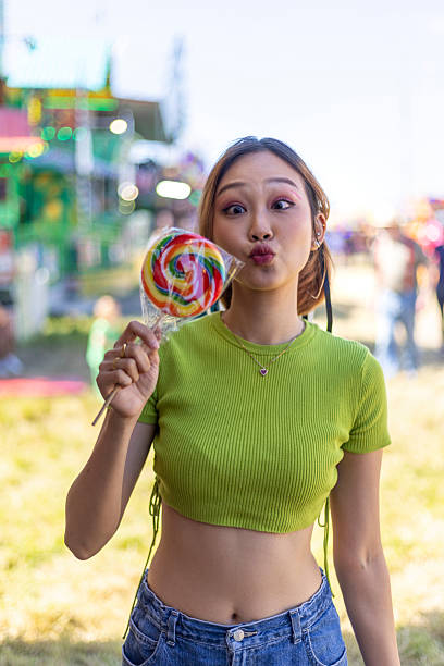 Three-quarter-length portrait of a young adult female having fun at a fairground in Newcastle-upon-Tyne, North East England. She is holding a multicoloured giant lollipop, making a cross-eyed face towards the camera. It is a summers day and she is wearing casual clothing.Videos similar to this scenario available.