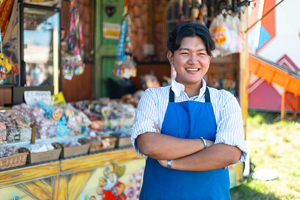 Waist-up portrait of a young adult male worker at a fairground in Newcastle-upon-Tyne, North East England. He is smiling with his arms folded, wearing an apron and standing in front of a colourful confectionary stall. It is a sunny summers day and he is wearing a pinstripe shirt beneath his apron.\n\nVideos similar to this scenario available.