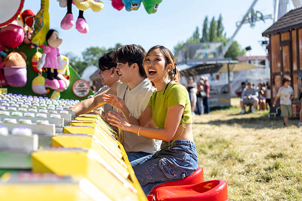 Side-view shot of three friends having fun at a fairground in Newcastle-upon-Tyne, North East England. They are all smiling as they play on the amusement arcades. It is a sunny day and they are wearing casual clothing. Fairground rides are visible in the background.Videos similar to this scenario available.