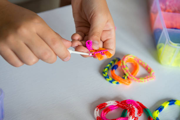 Child crafting colorful loom bands with hook on white table.