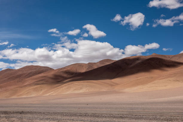 Arid desert hills under a vibrant blue sky with scattered clouds. Expansive sandy terrain reflecting sunlight. Rugged mountain landscape for travel and nature photography.