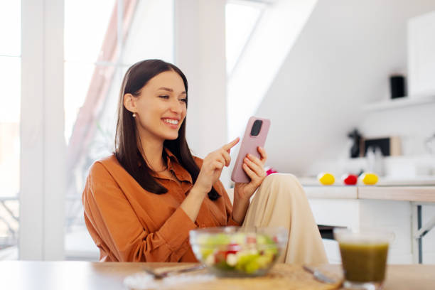 istock Glad positive lady chatting on cellphone in kitchen interior, sitting at table with bowl of fresh vegetable salad. Diet app, weight loss blog 2201055589