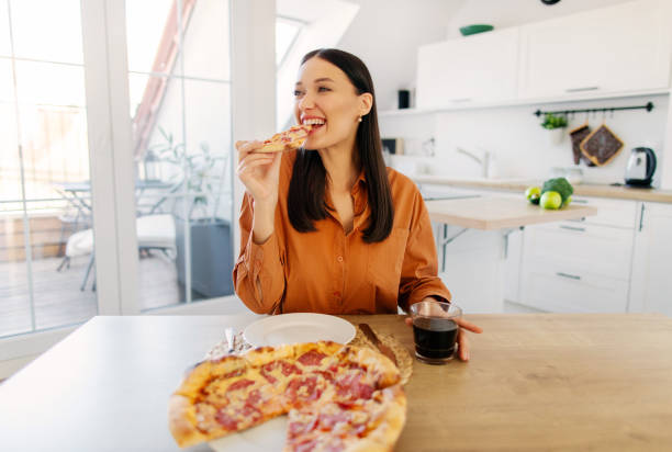 istock Satisfied lady enjoying homemade pizza, holding and biting tasty slice while sitting at table in kitchen. Junk food lover eating Italian pizza 2201055865