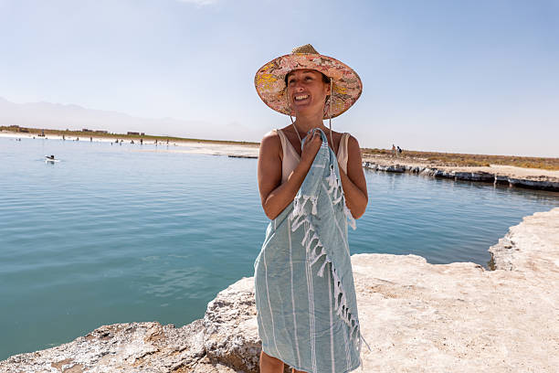 Woman in sun hat enjoying a sunny day near a calm lake. Content outdoor moment wrapped in a striped towel near water. Relaxation and leisure by the lakeside.