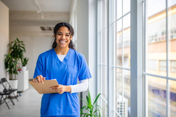 istock Smiling nurse in scrubs holds clipboard while standing in a bright hospital hallway with large windows 2201087697