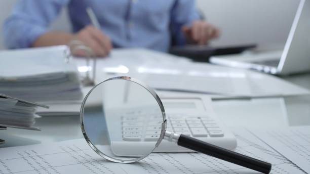 istock Magnifying glass, calculator, pen and documents are lying on accountant's desk. Businessman wearing blue shirt is working with documents on the background. Business people, audit concept 2201097886