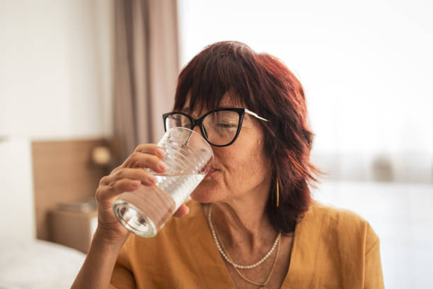 istock Mature woman wearing glasses drinking water while relaxing indoors 2201098243