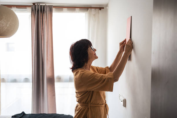 A middle-aged woman in casual clothing adjusting a picture frame on a bedroom wall. Enhancing home decor with personal touch.