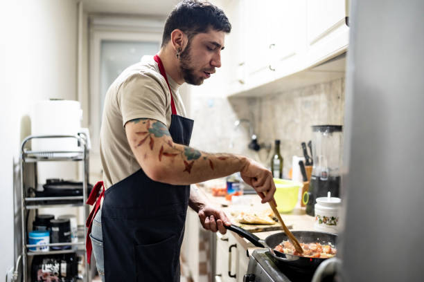 istock Young Male Cooking in Kitchen Wearing Apron While Stirring Food in Pan 2201109647