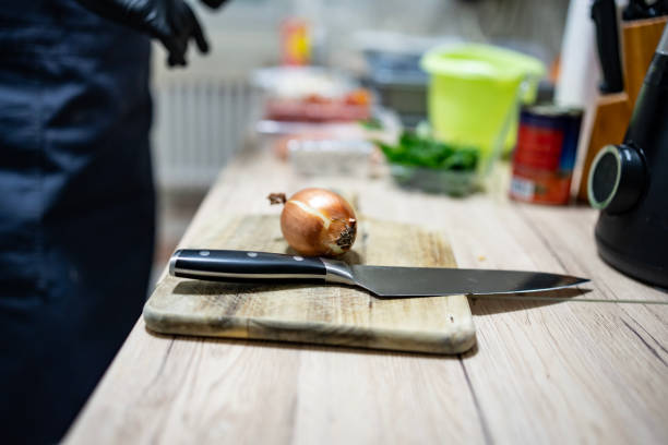 A professional kitchen setup displaying a fresh onion on a cutting board with a sharp knife, a person in the background, and various ingredients prepared for cooking.