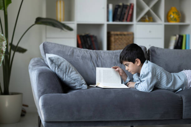 istock Boy lying on the sofa at home in day reading a book 2201121439