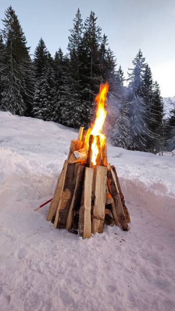 istock A bonfire burning in a snowy landscape, with logs arranged in a teepee shape. Flames rise high, emitting smoke. Snow-covered trees and mountains form a scenic backdrop. 2201131529