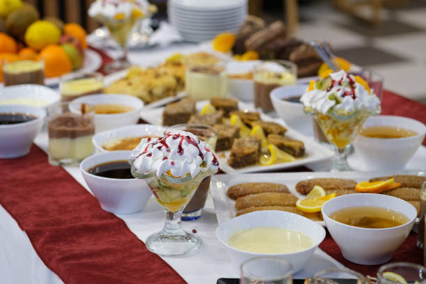 A table laden with an assortment of sweet treats, including fruit tarts, cakes, pastries, and creamy desserts.  Various beverages, including tea and coffee, complement the delectable selection. The setting suggests a buffet or catered event.