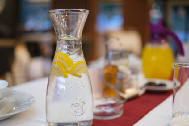 A clear glass pitcher filled with water and lemon slices sits on a table.  The background is blurred, showing a dining setting with other glassware and possibly a breakfast spread. The scene is bright and suggests a casual, healthy setting.
