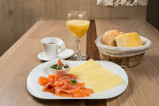 A breakfast arrangement features salmon, cheese slices, rolls in a basket, coffee, and orange juice on a wooden table, creating a fresh and inviting scene.