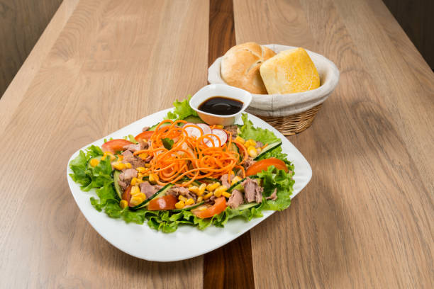 A vibrant salad with lettuce, tomatoes, cucumbers, carrots, corn, and a protein source, served with a dipping sauce and a side of bread rolls. It is on a wooden table.
