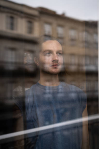 istock Pensive long-haired male with earring staring through window, city silhouette reflecting, mind wandering amid urban landscape background 2201157425