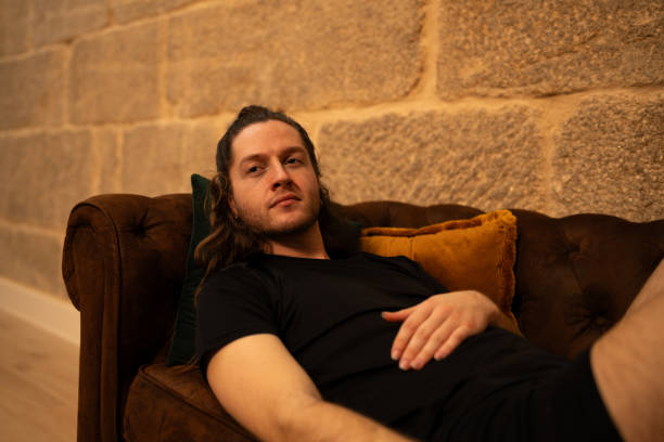 istock Bearded male reclining on vintage leather sofa, surrounded by rustic stone wall, wearing black tee and enjoying calm, serene atmosphere 2201157682