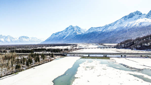 A drone shot shows the Knik Arm Bridge located in Alaska. This bridge is a major bridge for those traveling from Anchorage into other areas north of the city. A railroad bridge can be seen in the distance