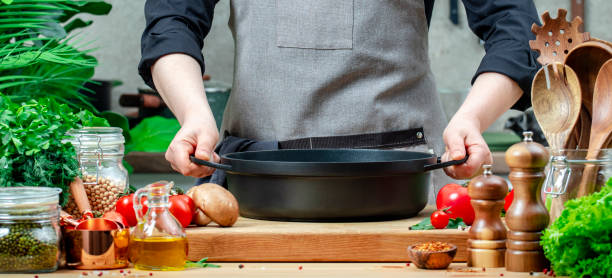 istock Home cook holds cast iron saucepan in hands. Cozy kitchen with wooden table, kitchenware, vegetables, herbs and ingredients for cooking. Healthy eating, culinary, recipes, food blogging 2201167404