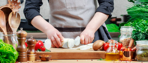 istock Home chef cuts white radish with santoke knife. Cozy kitchen with wooden table, kitchenware, vegetables, herbs and ingredients for cooking. Healthy vegan eating, culinary, recipes, blogging 2201167412