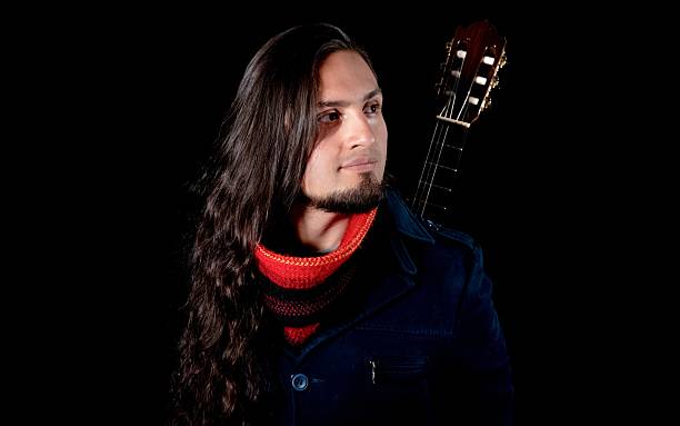 Portrait of a musician with long hair and a beard, wearing a red scarf and dark coat, posing with a classical guitar against a black background.