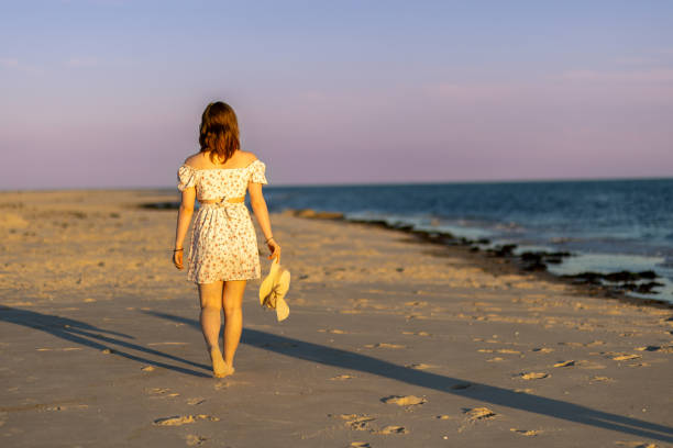A young woman strolls along the sandy beach at sunset, enjoying the serene atmosphere. She carries a light sweater and gazes at the tranquil ocean waves.