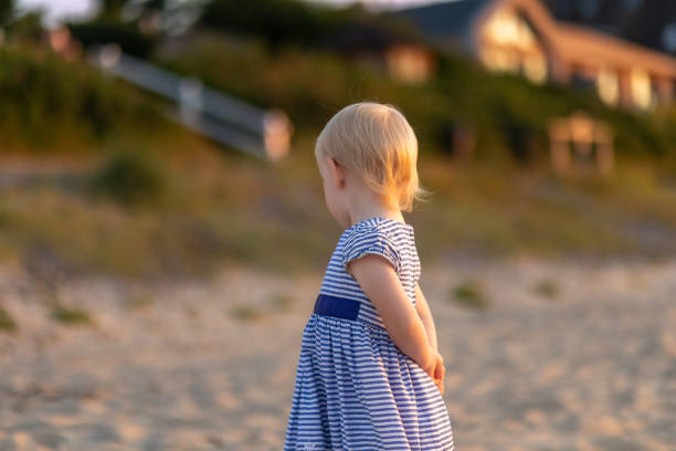 istock Child exploring the sandy beach at sunset while wearing a striped dress and looking away from the camera 2201182786