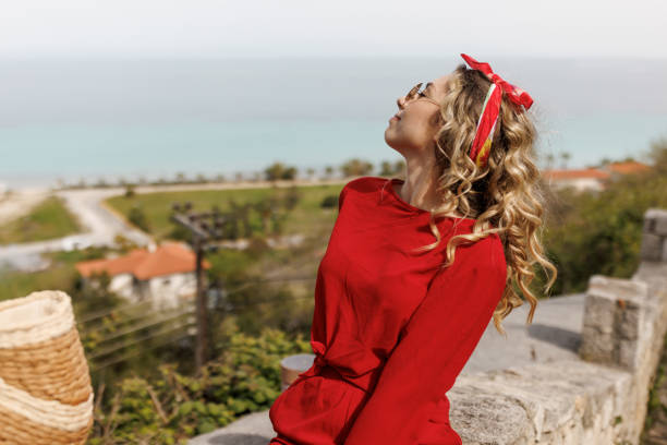 Beautiful young woman in red clothes sitting on stone wall and relaxing in old coastal town