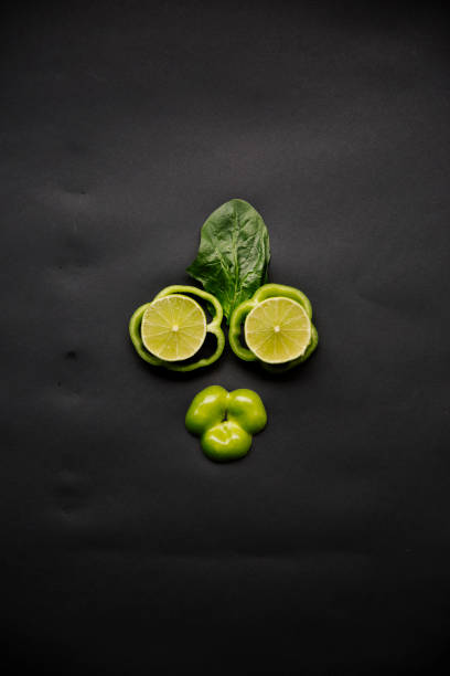 A top-down view of fresh green vegetables and a sliced lime arranged on a dark surface. The composition includes broccoli florets, green chili peppers, cucumber slices, and bell peppers, emphasizing organic, natural ingredients for healthy cooking. This vibrant image is perfect for themes related to nutrition, fresh produce, vegetarian meals, Thai cuisine, farm-to-table concepts, and food photography.