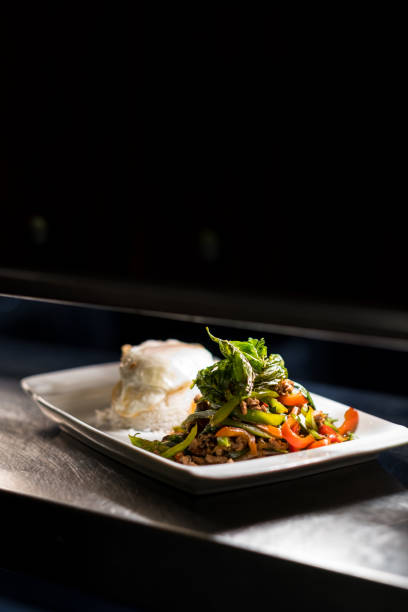 A delicious and spicy Thai Pad Krapow Moo Sap, featuring stir-fried minced pork with holy basil, chili, and garlic, served with a perfectly cooked fried egg on a white plate. This classic Thai street food dish is a staple in Thai cuisine, offering a bold combination of flavors with a savory, spicy, and aromatic taste. Captured in natural lighting on a rustic wooden table, this dish is a popular choice among food lovers and perfect for Thai restaurant menus, food blogs, and culinary photography.