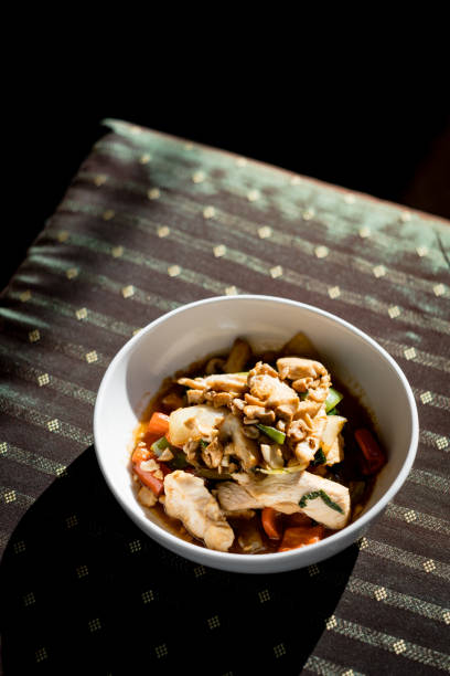 A beautifully plated Thai-style chicken and vegetable stir-fry, featuring tender chicken pieces, crisp vegetables, and a flavorful sauce, topped with crushed peanuts for added texture. Served in a ceramic bowl on a rustic wooden table, the dish is illuminated by dramatic lighting, creating a warm and inviting atmosphere. This image captures the essence of authentic Thai cuisine, perfect for food lovers and culinary enthusiasts.