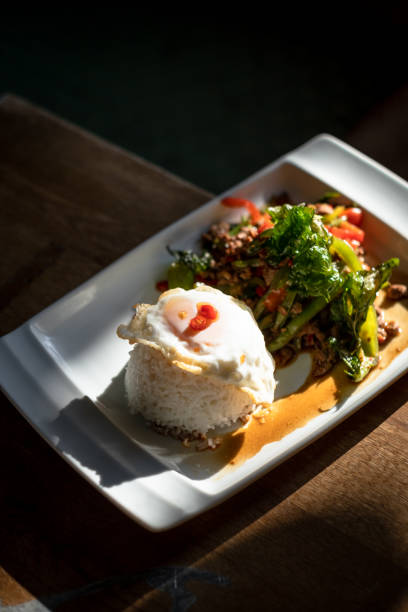A delicious and spicy Thai Pad Krapow Moo Sap, featuring stir-fried minced pork with holy basil, chili, and garlic, served with a perfectly cooked fried egg on a white plate. This classic Thai street food dish is a staple in Thai cuisine, offering a bold combination of flavors with a savory, spicy, and aromatic taste. Captured in natural lighting on a rustic wooden table, this dish is a popular choice among food lovers and perfect for Thai restaurant menus, food blogs, and culinary photography.