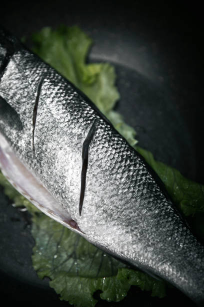 A detailed close-up shot of a fresh raw fish with shiny silver scales, featuring deep knife cuts for seasoning or marination. The fish is placed on a dark slate surface with green lettuce leaves, highlighting its freshness and texture. Ideal for seafood restaurants, culinary presentations, fish market promotions, healthy diet concepts, and recipe content related to grilled or pan-fried fish dishes.