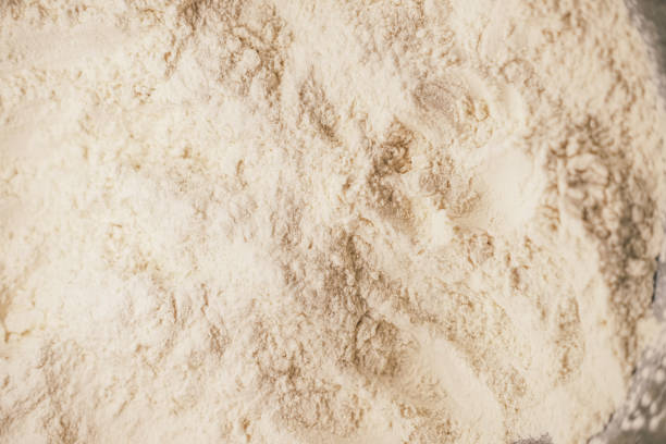 A detailed close-up view of white flour scattered across a flat surface, showcasing its fine powdery texture and natural patterns. The uneven distribution of flour creates an organic and rustic feel, emphasizing its role as a fundamental ingredient in baking and cooking. The soft lighting enhances the details, making this image ideal for food photography, culinary branding, and background textures for bakery and kitchen-themed designs.