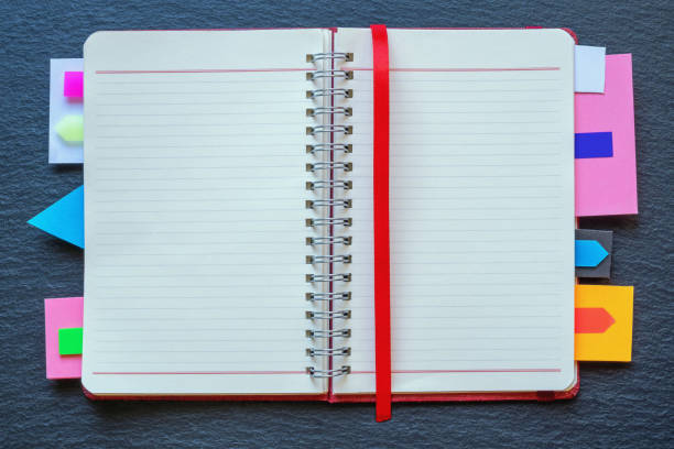 Open spiral bound notebook with bright colorful bookmarks on dark table. Flat lay, copy space