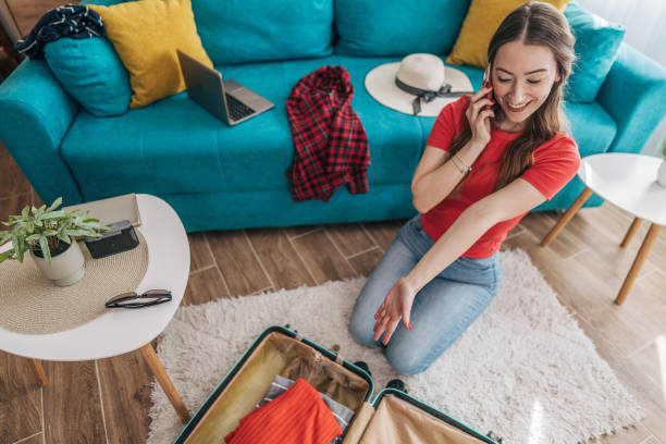 istock Young woman packing a suitcase while talking on smartphone at home 2201238008