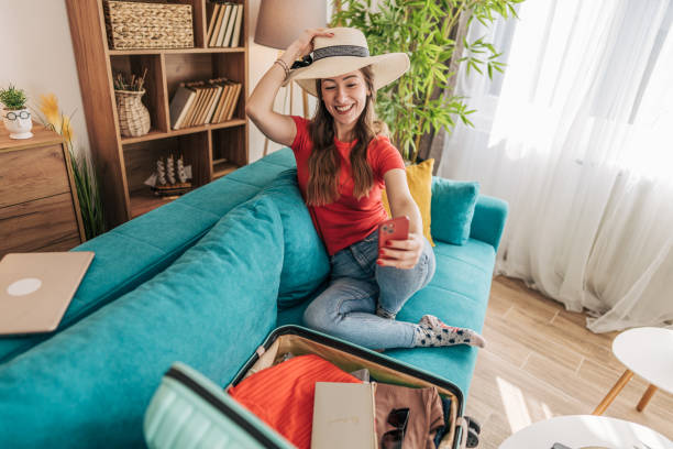 istock Young woman taking selfie while packing suitcase for summer vacation 2201239059