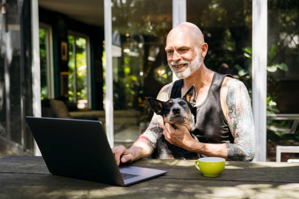 istock Cheerful senior man with tattoos using laptop and working outdoors, with Blue Heeler on lap 2201239385