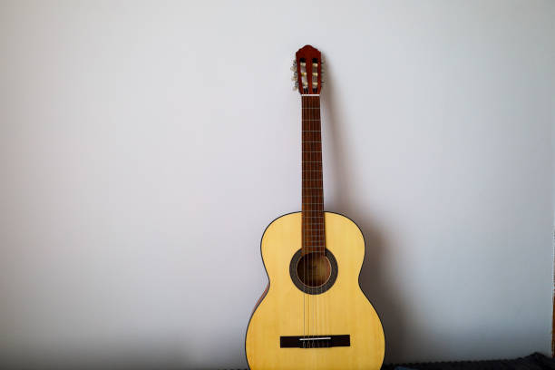 istock A classical acoustic guitar hangs on a light gray wall. The guitar is natural wood colored with a brown hue. The guitar neck is brown, with metal frets. 2201246487