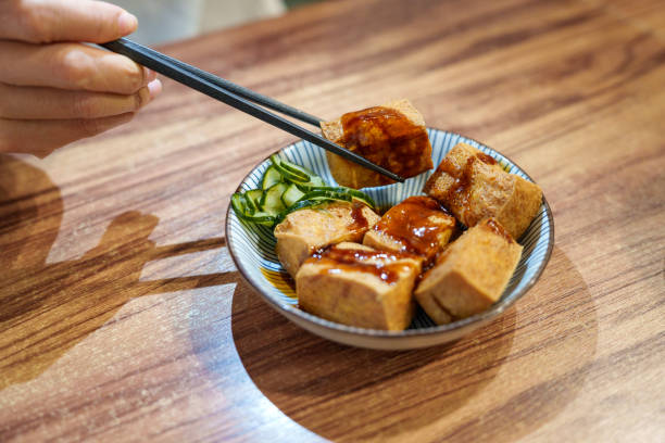 A close-up shot of delicious Taiwanese fried tofu with savory sauce and pickled cucumbers, served in a ceramic bowl. A diner uses chopsticks to enjoy this popular dish.