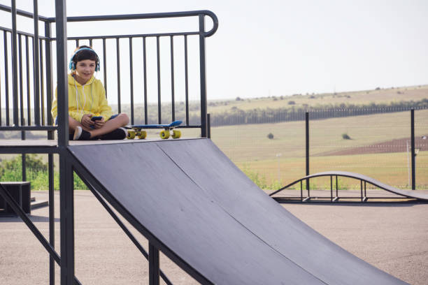 A smiling boy sits on a skate ramp with headphones on, engaged in a moment of leisure and music, surrounded by an open skatepark and green scenic views.